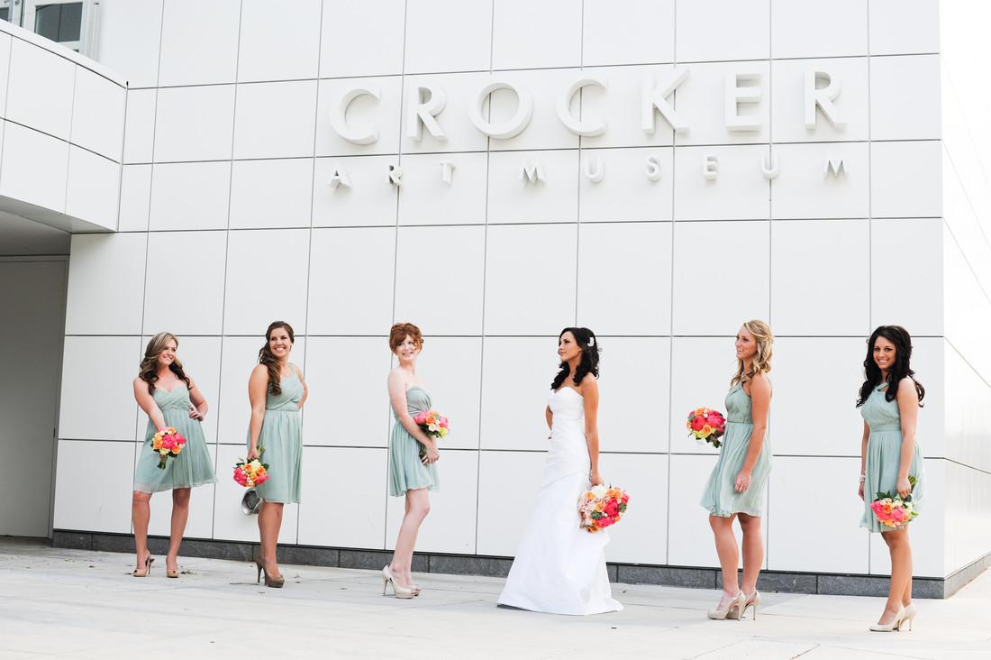 A photo of a bride in a white, sleeveless bridal fown holding a colorful bouquet surrounded by 5 bridesmaids in short, silver dresses. They are standing underneath the Crocker Art Museum side outside the front entrance of the Museum.