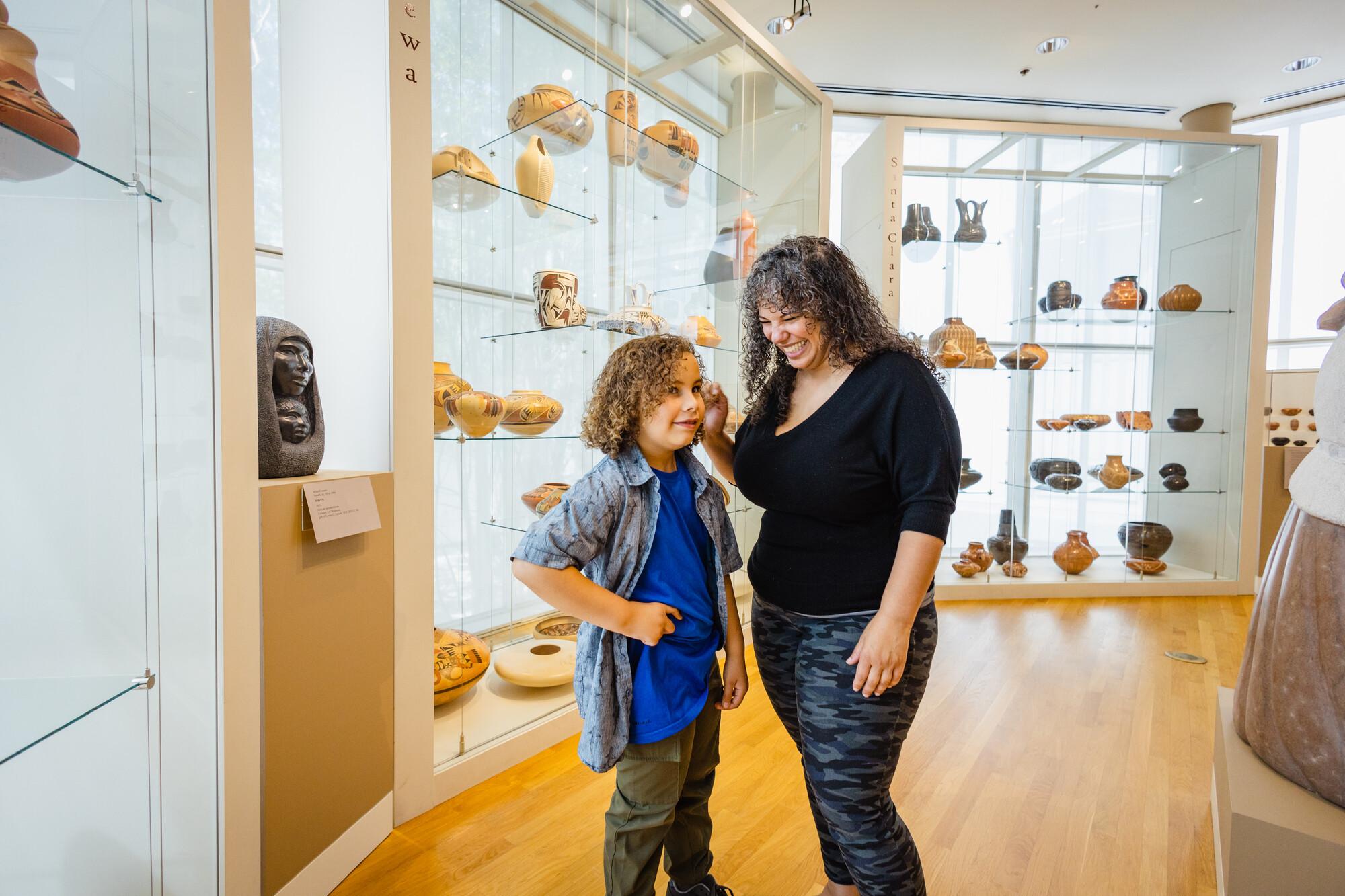 A mother with shoulder-length curly grey hair wearing a black shirt and dark pants stands in one of the Crocker's galleries laughing. A child with chin length curly hair wearing a light blue collared shirt over a bright blue shirt and pants, stands next to her, also laughing.