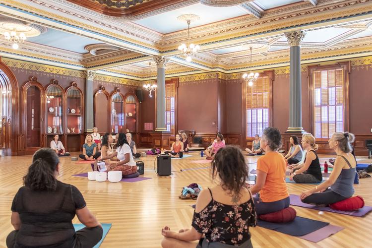 A group of people sit in the Crocker's historic ballroom on yoga mats. In the middle, a person leads a sound healing class with white bowls.