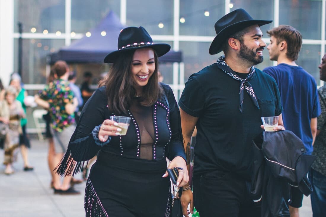 A man and woman dressed in black Western wear stroll through the Crocker's outdoor courtyard laughing with drinks in their hands.
