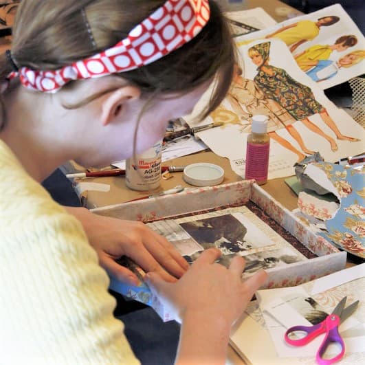 A person wearing a headband and pale yellow sweater is working on a mixed-media assemblage work.